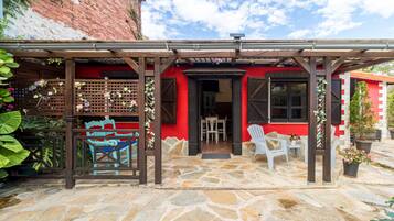 Un patio empedrado con un pérgola de madera, un edificio rojo con una puerta negra y sillas de plástico blanco.