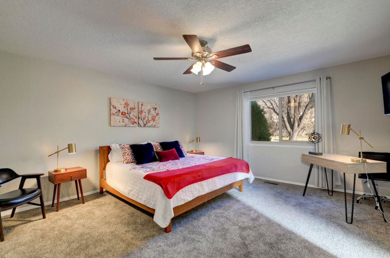 A bedroom with a bed, a desk, a chair, a ceiling fan, and a window.