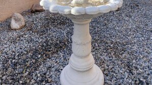 Ein weißer Steinbrunnen mit mehreren Stufen, von denen Wasser herabfließt, auf einem Kiesgrund, im Hintergrund ein Gebäude und Bäume.