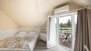 A bedroom with a bed, a radiator, a window, and an open door leading to a balcony with a table and chairs. - TópART Apartman I: Apartment with terrace (Balatonfenyves)