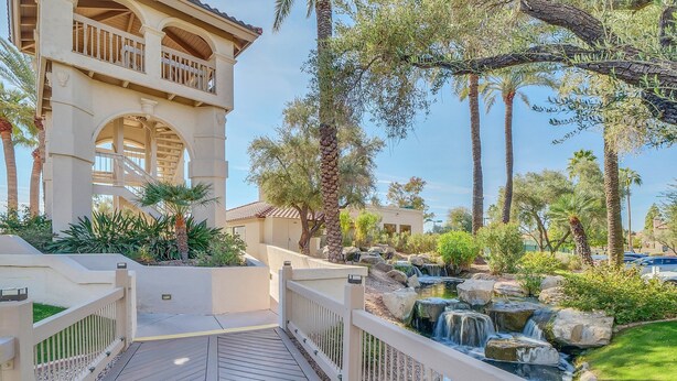 A wooden deck leading to a pool with a waterfall, surrounded by palm trees and a building with a balcony. - STUNNING Views, SUBERB Location..NEW NEW NEW to The Racquet Club in Scottsdale! (Scottsdale)