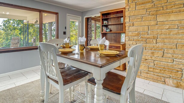 A dining area with a wooden table set for four, surrounded by white chairs. A stone wall and a window with a view of trees are visible. - Private Yard & Grill: Retreat Near Norfolk Lake! (Mountain Home)
