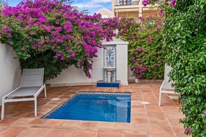 A swimming pool with two white lounge chairs, surrounded by a tiled patio and vibrant purple bougainvillea flowers.