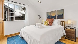 Une chambre à coucher avec un grand lit, une table de chevet avec une lampe, une fenêtre avec des rideaux et un tableau accroché au mur.
