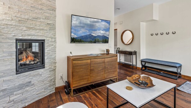 A modern living room with a stone fireplace, a flat-screen TV, a wooden cabinet, a bench, and a mirror on the wall. - NAU/DT | Elevated 2 bdrm | AC | Dog Friendly (Flagstaff)