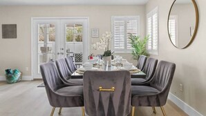 Een eetruimte met een ronde tafel, twee grijze fauteuils met gouden details, een kroonluchter en een grote spiegel aan de muur.