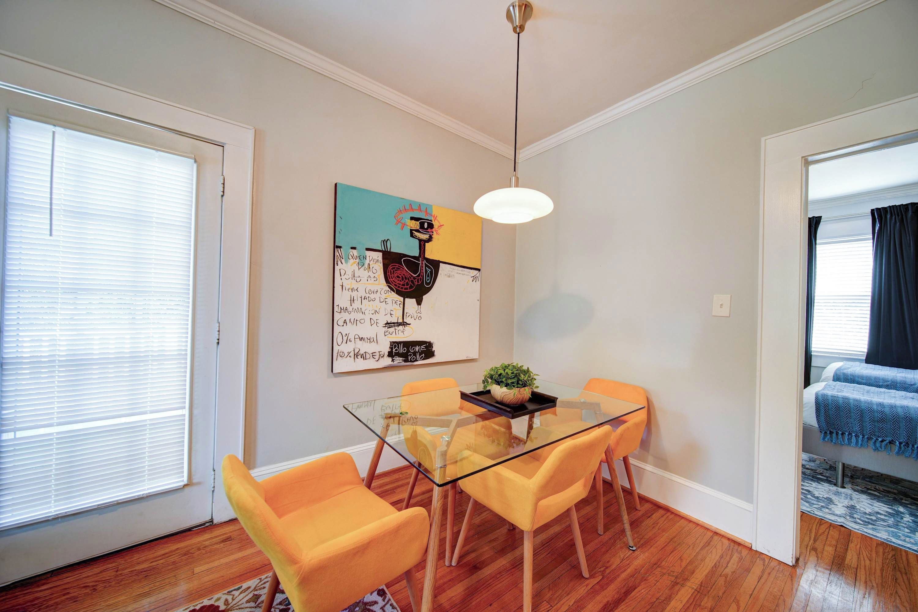A dining area with a glass table, orange chairs, a ceiling light, and a wall with a colorful painting.