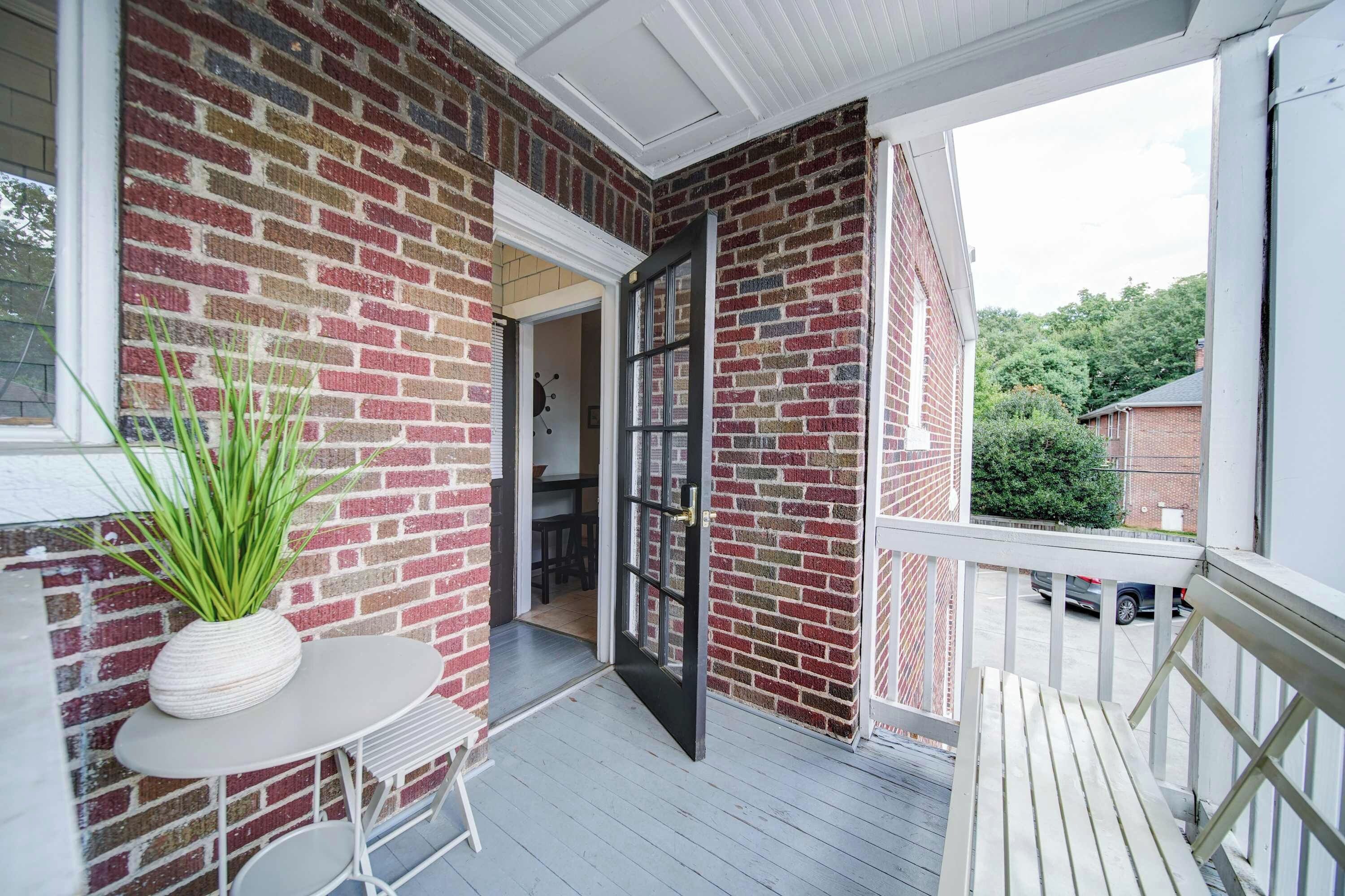 A covered porch with a brick wall, a potted plant, a table, and a door leading to an interior space.