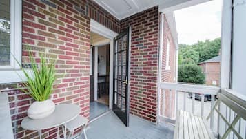 A covered porch with a brick wall, a potted plant, a table, and a door leading to an interior space.