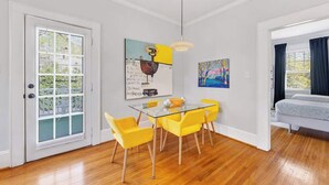 Un comedor con mesa de vidrio y sillas amarillas, un cuadro en la pared y una cama en la habitación contigua.