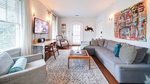 Ein Wohnzimmer mit grauem Sofa, Couchtisch und Fernseher auf einem Schrank. Eine Tür mit Stierverzierung ist in der Wand zu sehen, darüber hängt ein farbenfrohes abstraktes Gemälde.