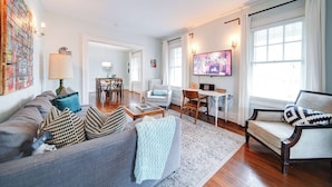 Ein Wohnzimmer mit grauem Sofa, einem Couchtisch und einem Fernseher. Es gibt eine Essecke mit Tisch und Stühlen.