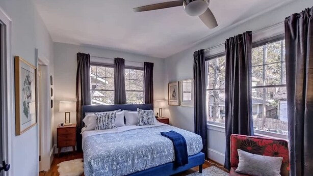 A bedroom with a bed, a chair, a ceiling fan, and large windows with curtains.
