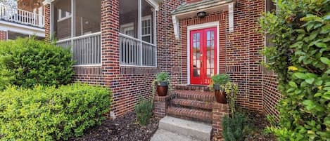 Une maison en briques avec une porte rouge, un porche couvert et un petit jardin devant la maison, agrémenté de plantes.