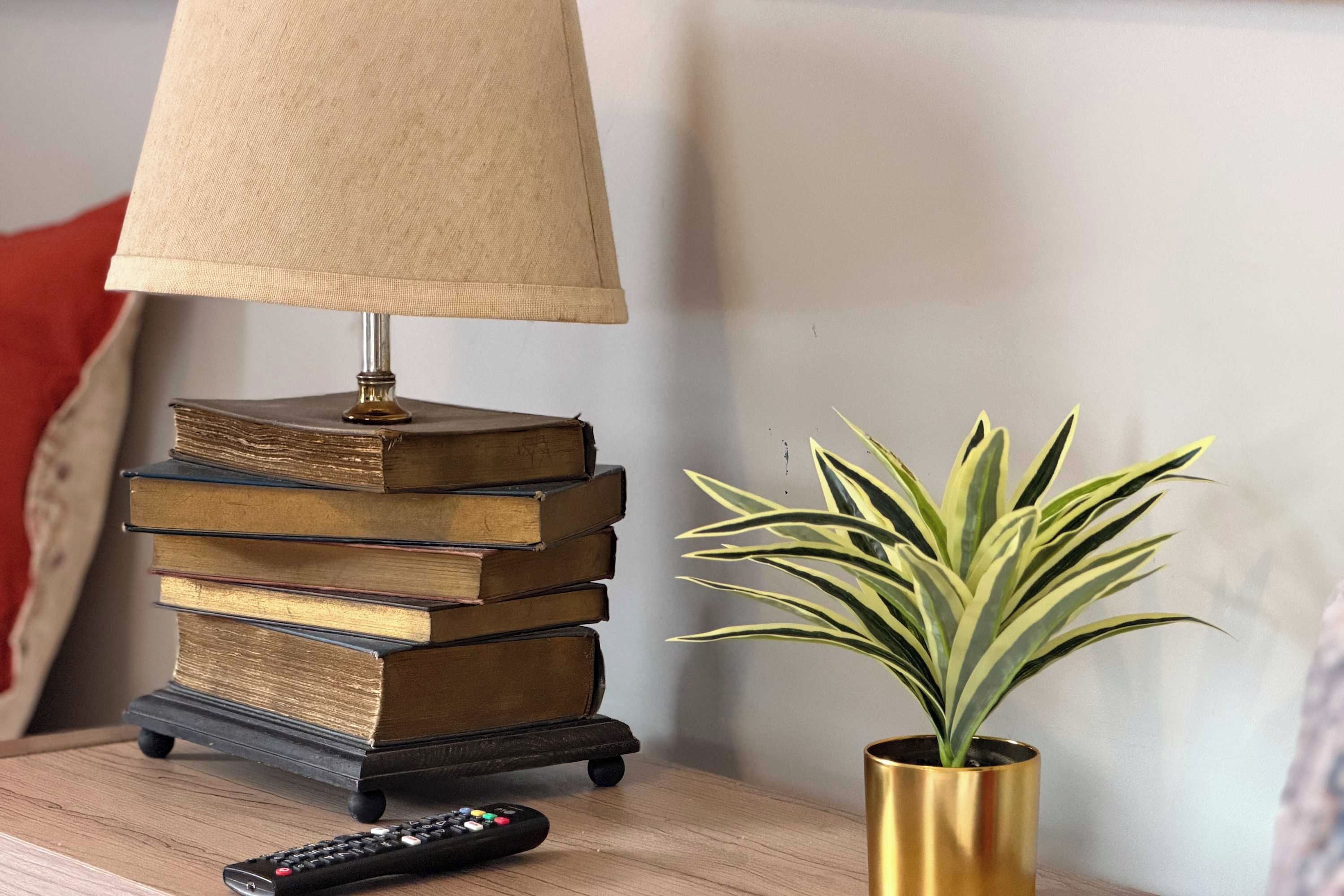 A stack of books on a small table with a lamp and a remote control beside it.