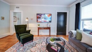 A living room with a green armchair, a coffee table with a glass top, a painting on the wall, and a door with a dark frame. - The Home Collection: Historic Step-Free 2BR, Pet Friendly & Close to  Atrium CMC (Charlotte)
