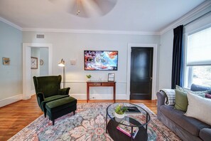 Ein Wohnzimmer mit einem grünen Sessel, einem Couchtisch mit Glasplatte, einem Gemälde an der Wand und einer Tür mit dunklem Rahmen.