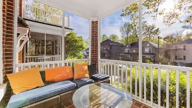Ein überdachter Balkon mit einem Glastisch und einer Rattan-Couch mit orangefarbenen Kissen.