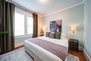 A bedroom with a large bed, a nightstand with a lamp, a painting on the wall, and a window with curtains. - The Home Collection: Pet-Friendly King Suite near South End & Freedom Park (Charlotte)