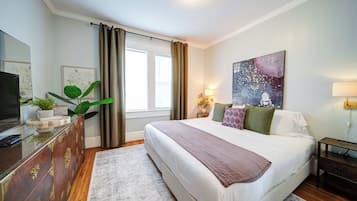 A bedroom with a bed, a dresser, a television, a painting, and a window with curtains.