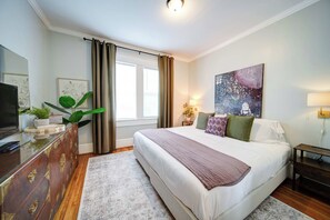 Un dormitorio con cama, cómoda, televisor, un cuadro y ventana con cortinas.
