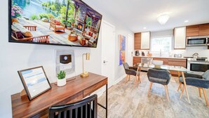 A modern kitchen with a large flat-screen TV, wooden cabinets, and a dining area with a glass table.
