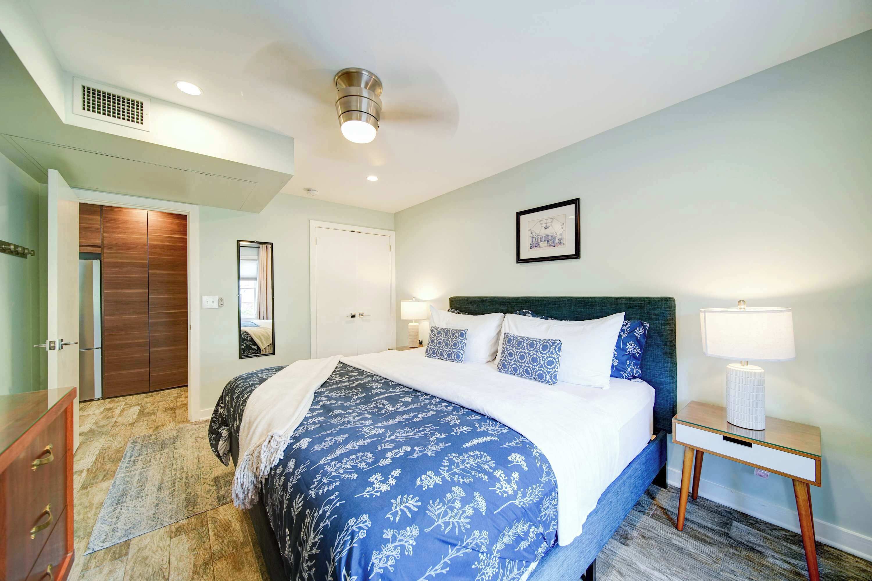 A bedroom with a bed, bedside lamps, a nightstand, a dresser, a door, and a ceiling vent.