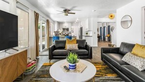 A modern living room with a flat-screen TV, a black leather sofa, a coffee table with a plant, and a ceiling fan. - Extravagant 4BD Home w/ Hot Tub & Pool Table (North Las Vegas)