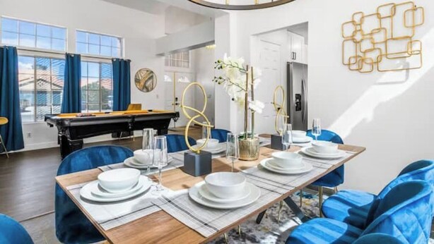 A dining area with a wooden table set for six, blue upholstered chairs, and a pool table in the background. - Extravagant 4BD Home w/ Hot Tub & Pool Table (North Las Vegas)