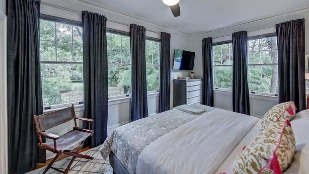 Un dormitorio con cama, una silla, un televisor y ventanas grandes con cortinas.