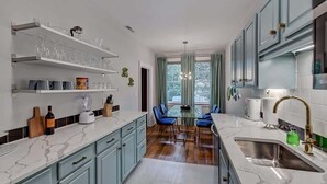 Una cocina moderna con encimeras de mármol, armarios azules, una zona de comedor con sillas azules y un ventanal con cortinas.