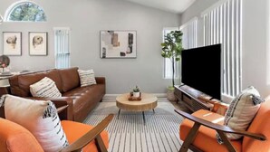 Ett modernt vardagsrum med en brun lädersoffa, orange fåtöljer, ett runt soffbord och en stor TV med platt skärm.
