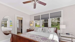A bedroom with a wooden bed, a ceiling fan, a large window with blinds, and a bedside table with a lamp. - Charming Condo Near Freedom Park | King Bed & Screened Porch (Charlotte)