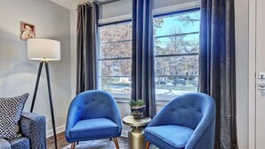 Une pièce avec deux fauteuils bleus, une lampe sur pied, une petite table avec une plante, et une vue sur une rue bordée d’arbres, visible à travers de grandes fenêtres.