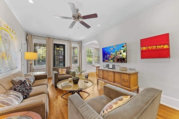 Bright living room in Charlotte featuring hardwood floors, large windows, comfortable seating, ceiling fan, and a mounted smart TV. Open and inviting space perfect for relaxing, entertaining, or unwinding after exploring the city.