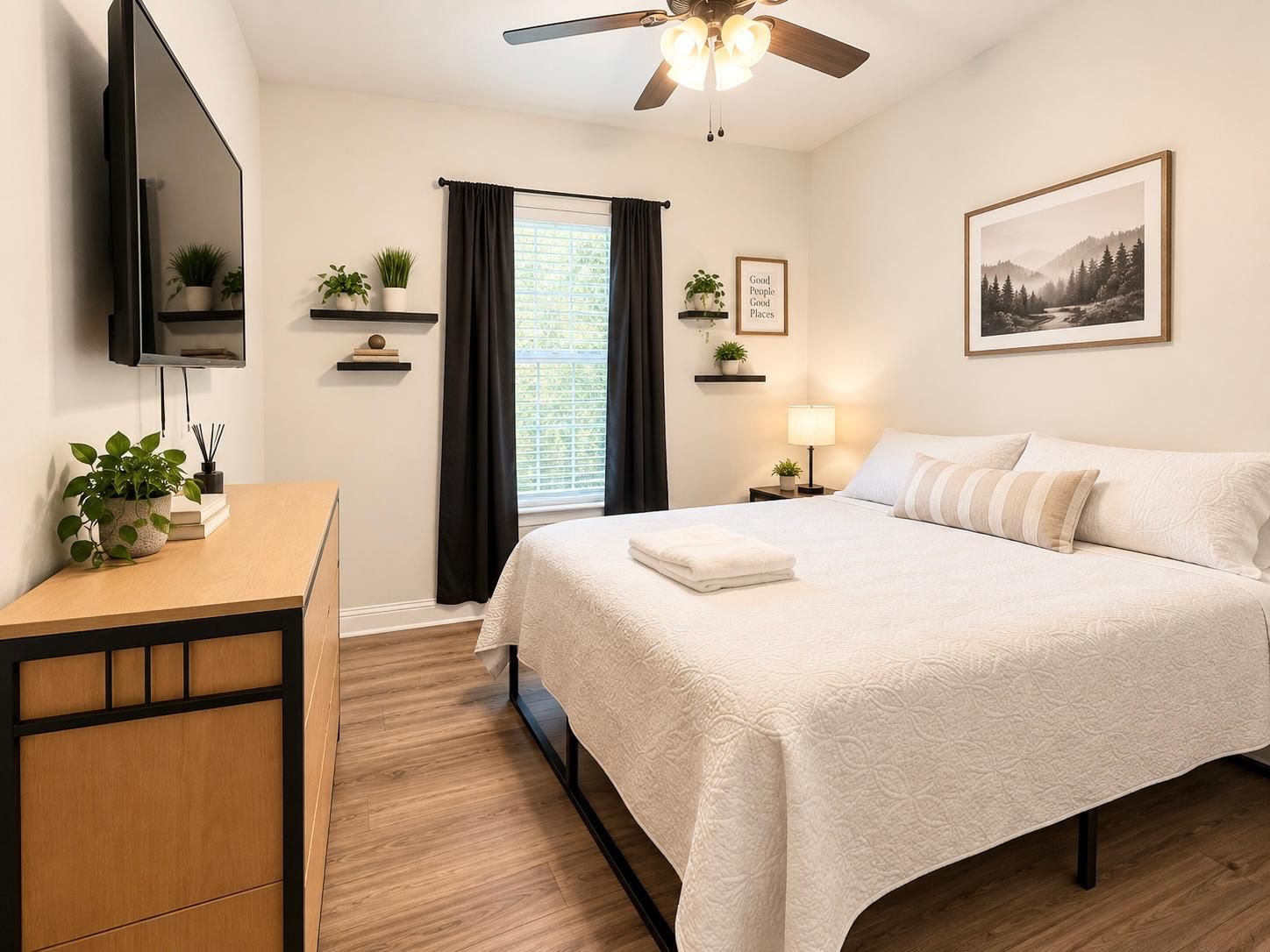 A hotel room with a bed, a desk, a TV, a ceiling fan, a window with curtains, and a framed picture on the wall.