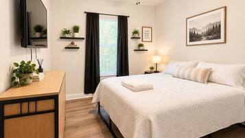 A hotel room with a bed, a desk, a TV, a ceiling fan, a window with curtains, and a framed picture on the wall.