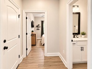 Un couloir avec des portes blanches, un plancher en bois et une salle de bain équipée d’un miroir, d’un lavabo et d’un porte-serviettes.