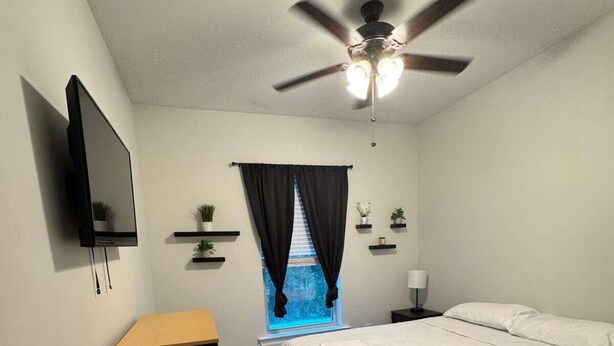 Una camera da letto con un letto, una TV, una ventola a soffitto, una finestra con tende e un comodino.
