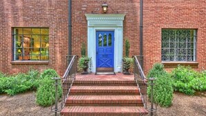 Un bâtiment en briques avec une porte bleue, des marches menant à l’entrée et un petit jardin agrémenté de plantes en pot.