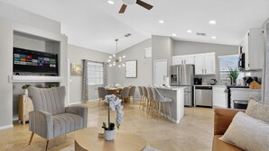 A modern living room with a flat-screen TV, grey upholstered armchair, round coffee table, and a dining area with a chandelier. - Spectacular 5BR Home: Pool and Relaxing Space! (North Las Vegas)