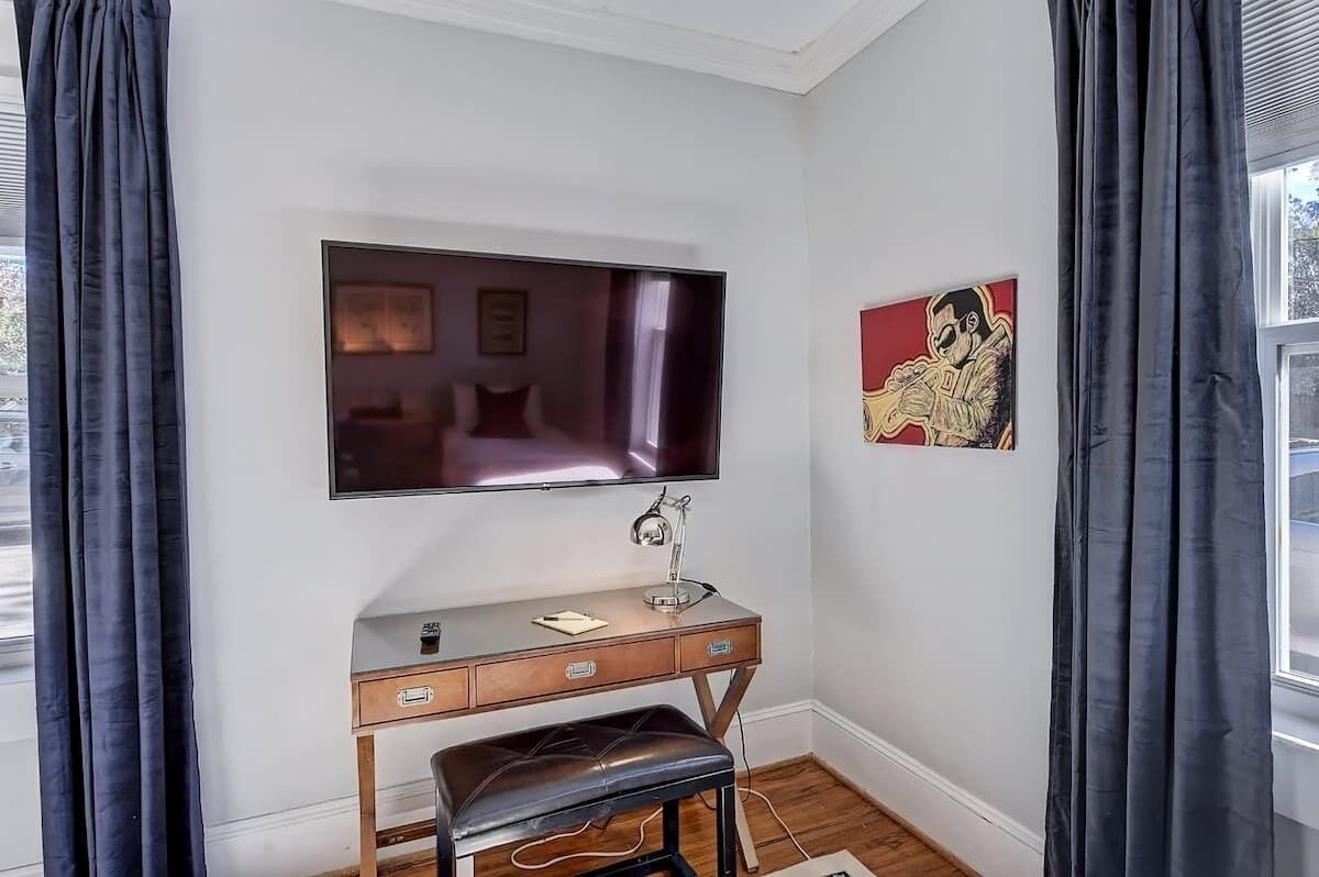 A bedroom with a flat-screen TV mounted above a wooden desk with drawers, a bench, and a small table. A painting is on the wall to the right of the TV.