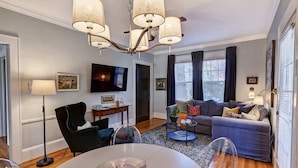 A modern living room with a grey sofa, a dining table, and a chandelier. - Historic Dilworth 3-Unit Stay | 6 King Beds + Parking | Near Atrium & Novant (Charlotte)