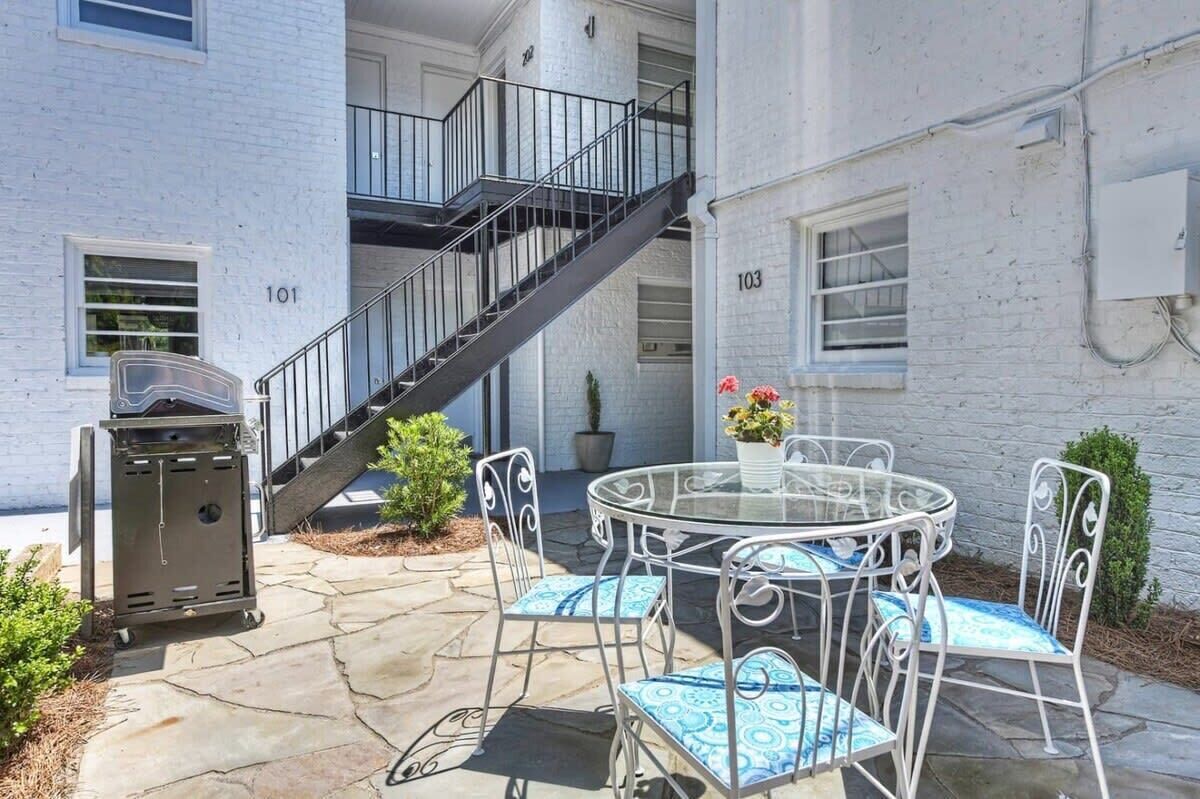 A patio area with a round glass table and metal chairs, a barbecue grill, and a staircase leading to an upper floor.