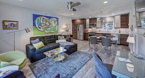 A modern living room with a sofa, coffee table, and chairs, a dining area with a table and chairs, and a kitchen with appliances and cabinets.