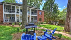 A two-story building with a porch, a barbecue grill, and blue Adirondack chairs in the yard.