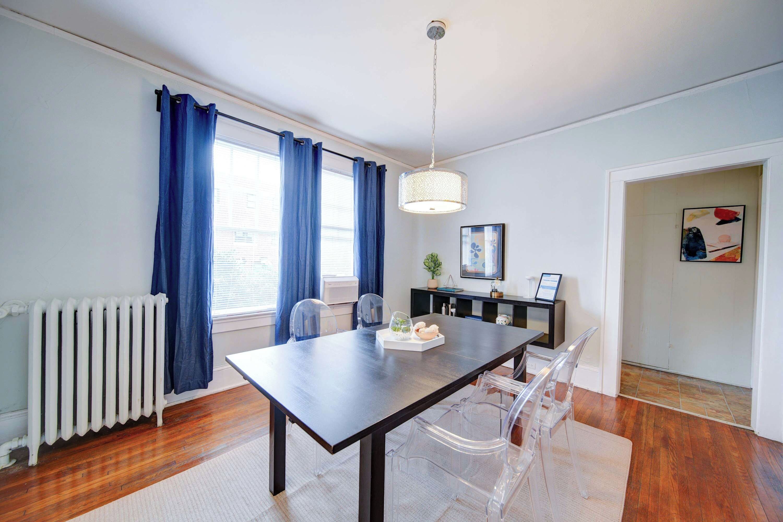 A dining area with a table, chairs, a hanging light fixture, a radiator, and a window with curtains.