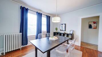 A dining area with a table, chairs, a hanging light fixture, a radiator, and a window with curtains.
