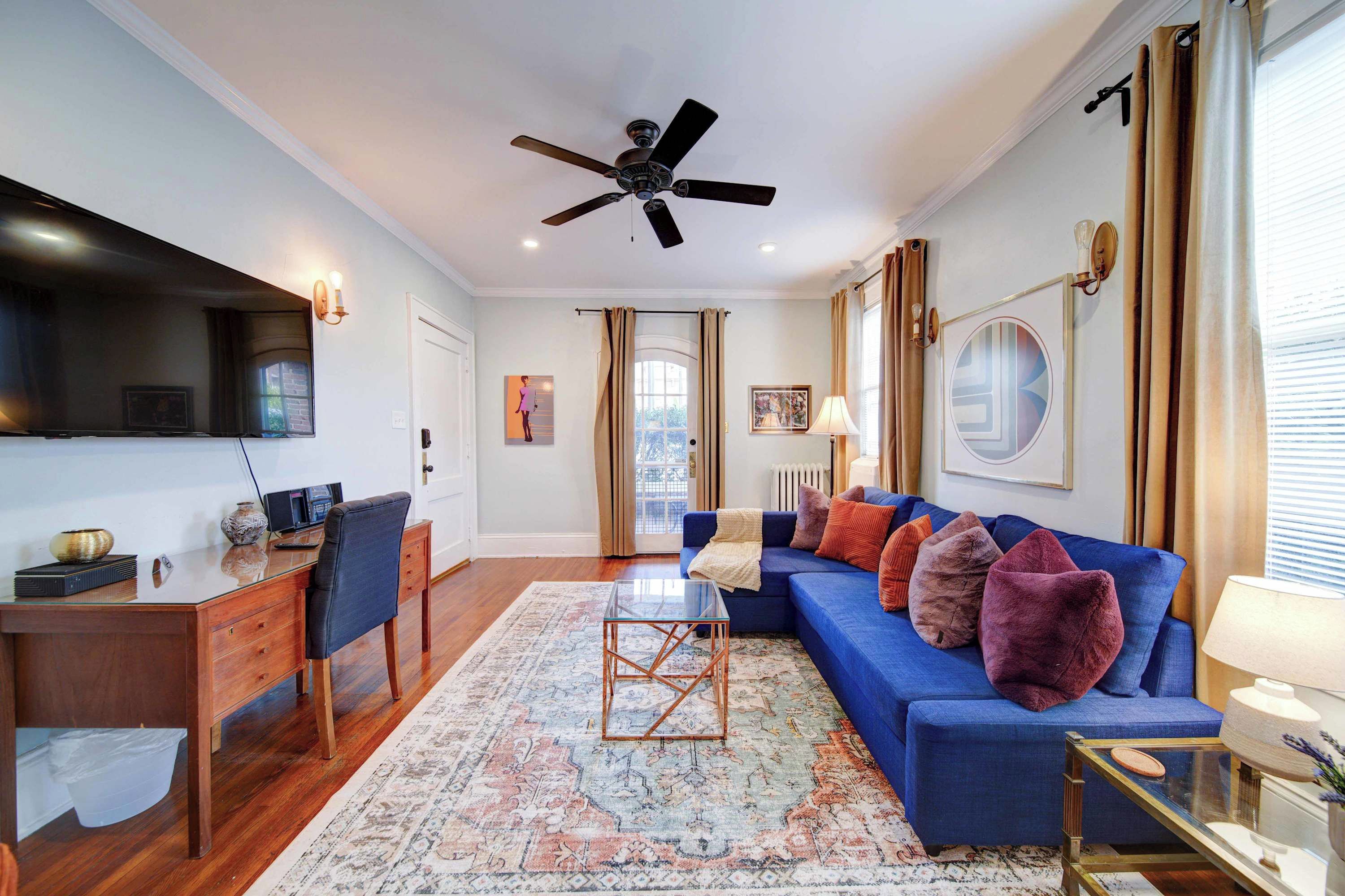 A living room with a blue sofa, a coffee table, a desk with a computer, a ceiling fan, and a large window with curtains.
