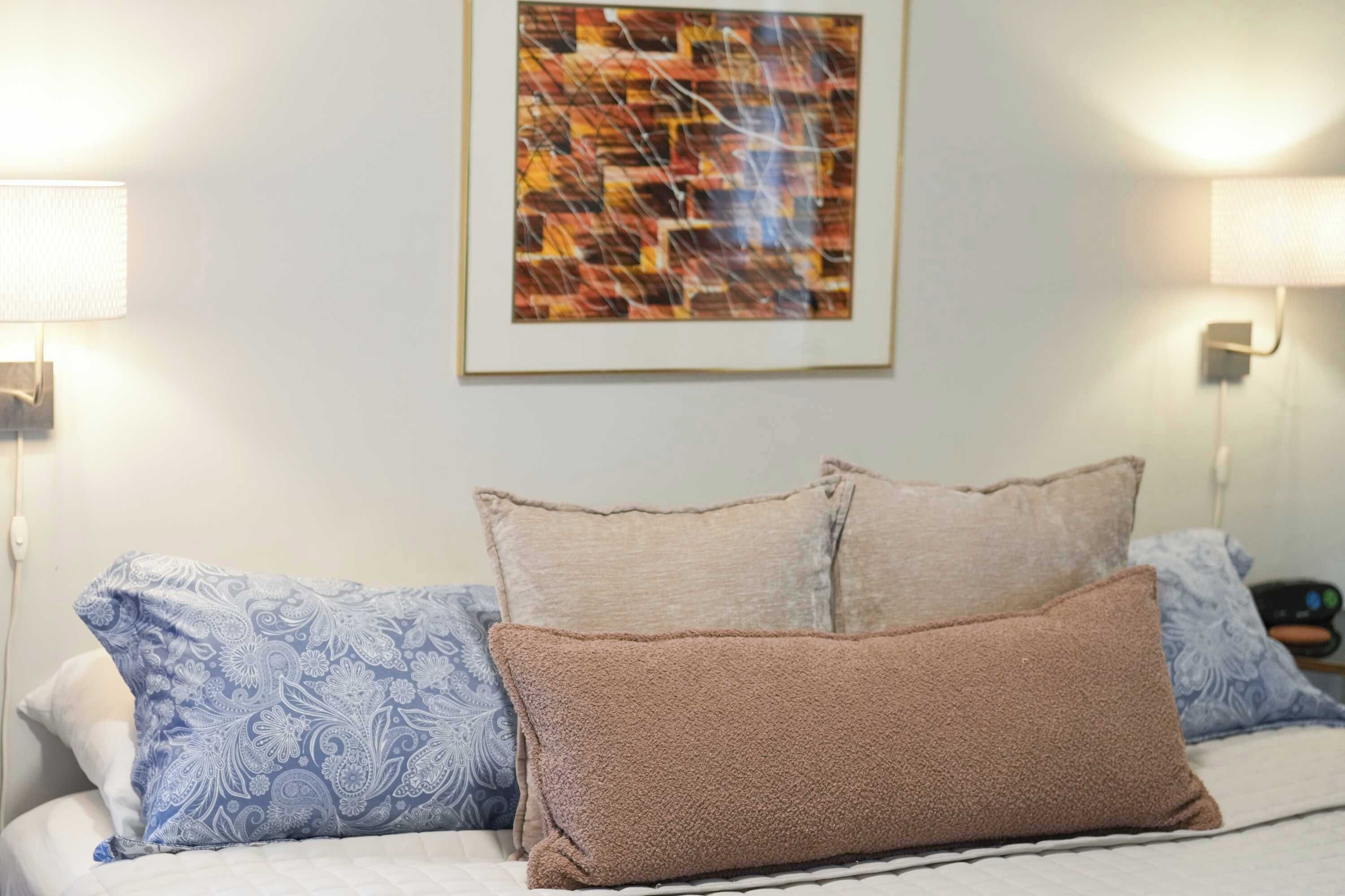A neatly made bed with patterned pillows, a framed abstract painting on the wall, and wall-mounted lamps.
